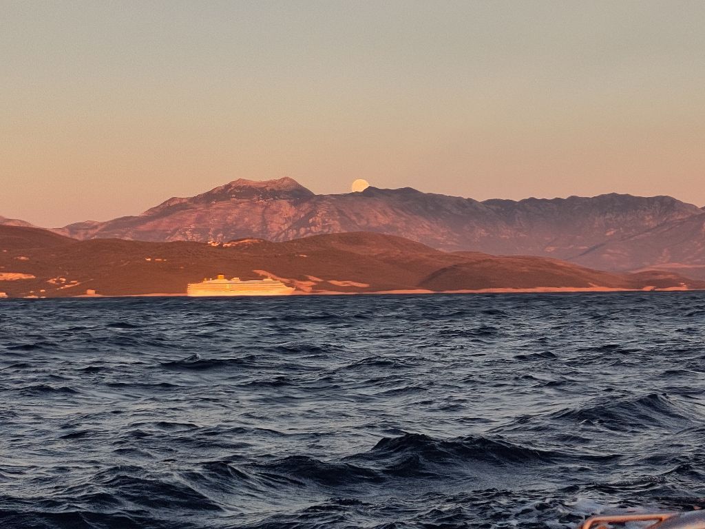 der Mond geht über dem Gebirge auf, darunter verlässt ein Kreuzfahrtschiff die Bucht